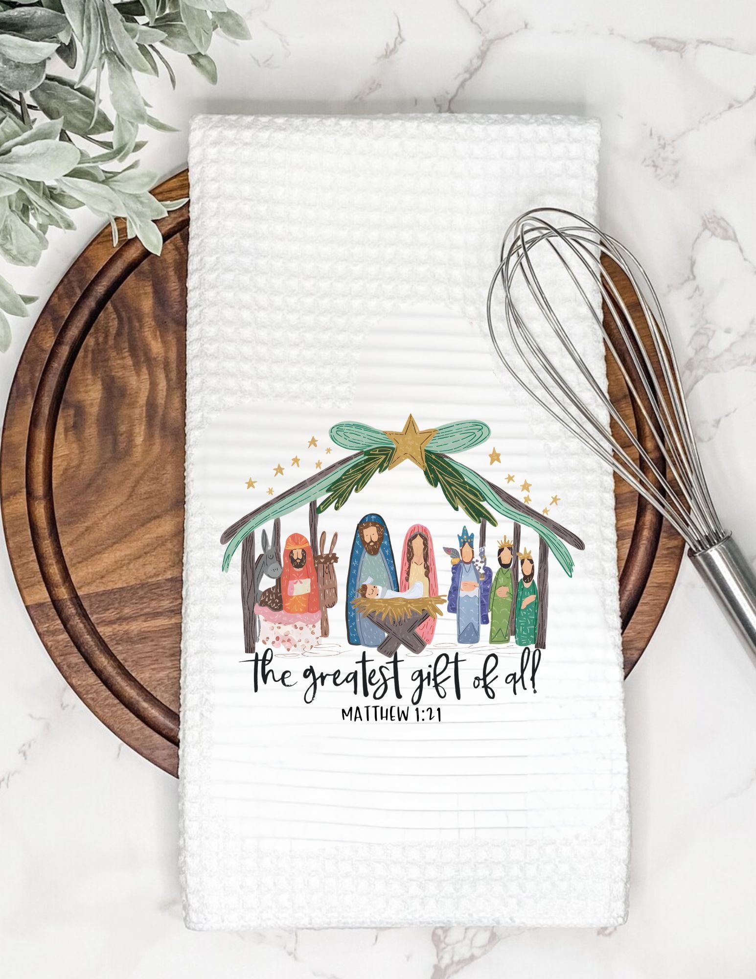 Napkin with nativity scene illustration and text on a marble surface with whisk and leaves.