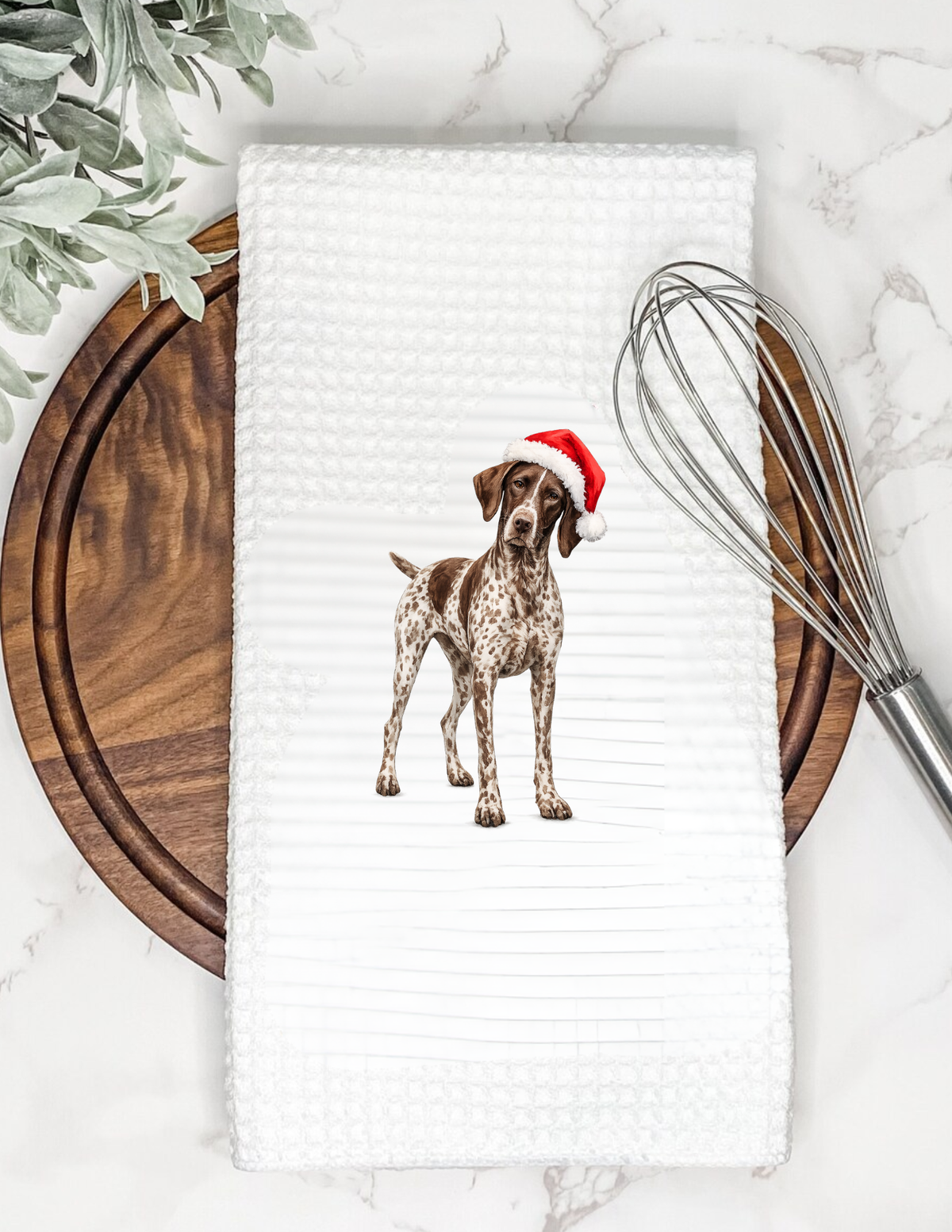 A white waffle-weave kitchen towel featuring a printed illustration of a brown and white spotted dog wearing a red Santa hat, styled on a marble surface with a wooden board, greenery, and a metal whisk.
