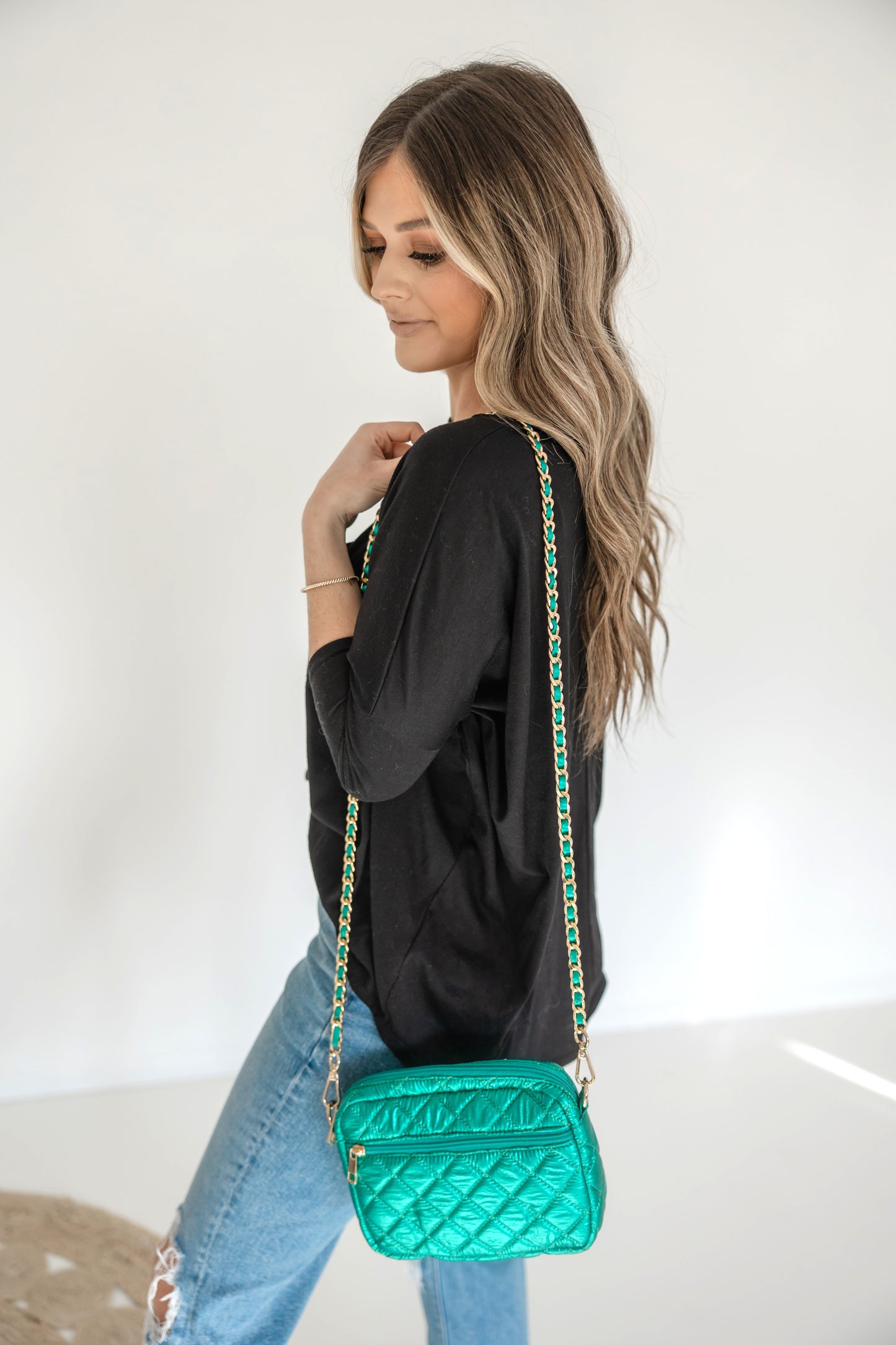 A woman wearing a denim shirt is showcasing a small quilted crossbody bag with a gold chain strap.