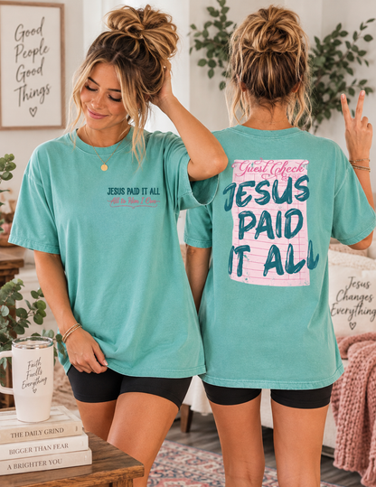 Two women modeling matching turquoise graphic T-shirts in a cozy indoor setting. The front view shows the text 'JESUS PAID IT ALL' in black with a pink heart outline, and the back view shows a large pink and blue graphic reading 'Jesus Paid It All'.