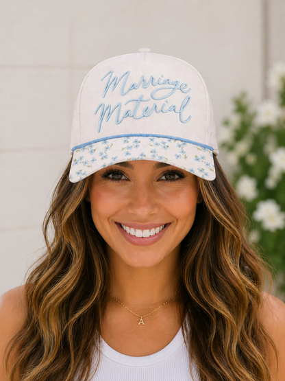 Woman wearing a white cap with 'Marriage Material' text and floral patterns, smiling outdoors.