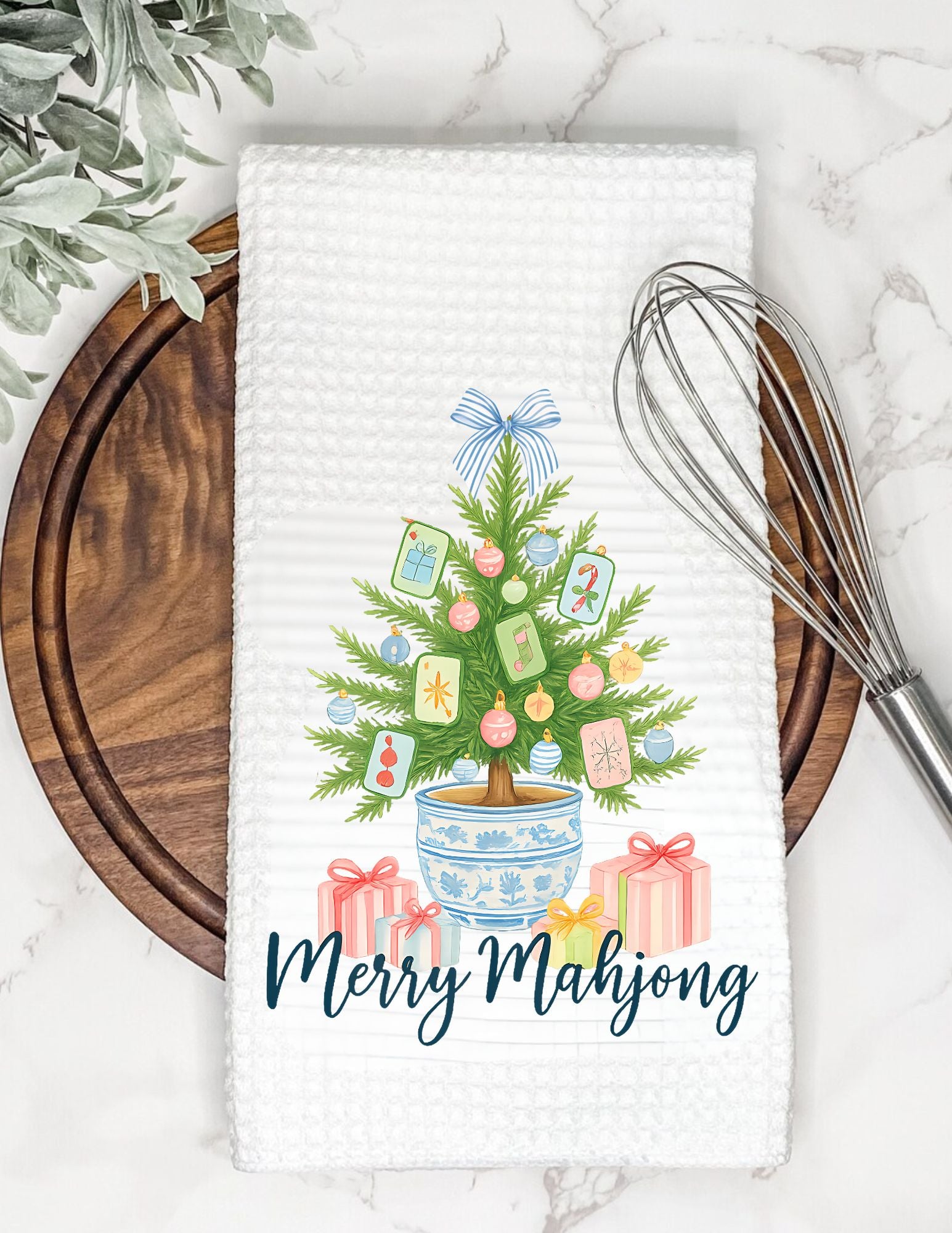 Decorative towel with a Christmas tree and presents design on a marble surface with a whisk and plant.