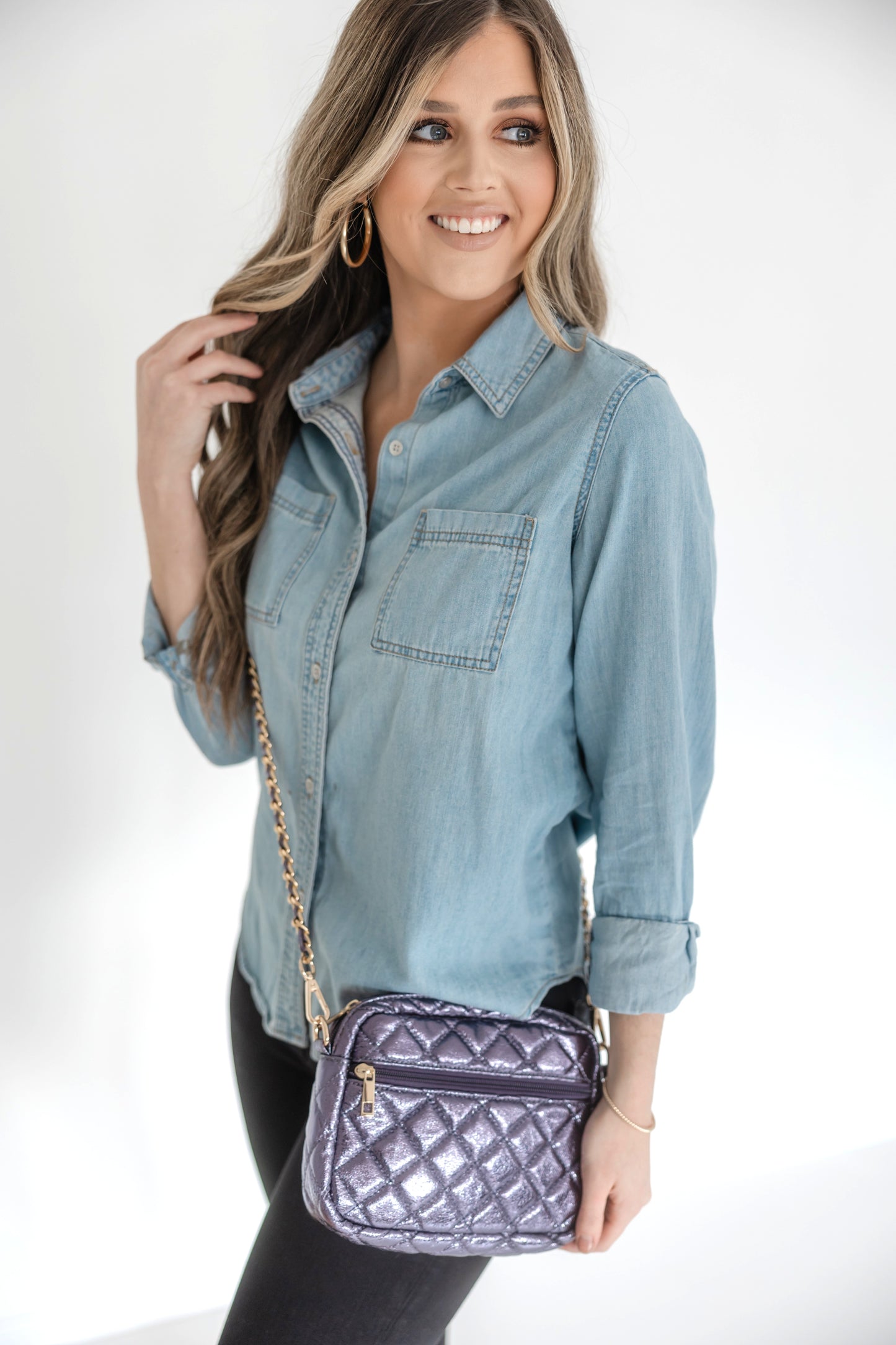 A woman wearing a denim shirt is showcasing a small quilted crossbody bag with a gold chain strap.