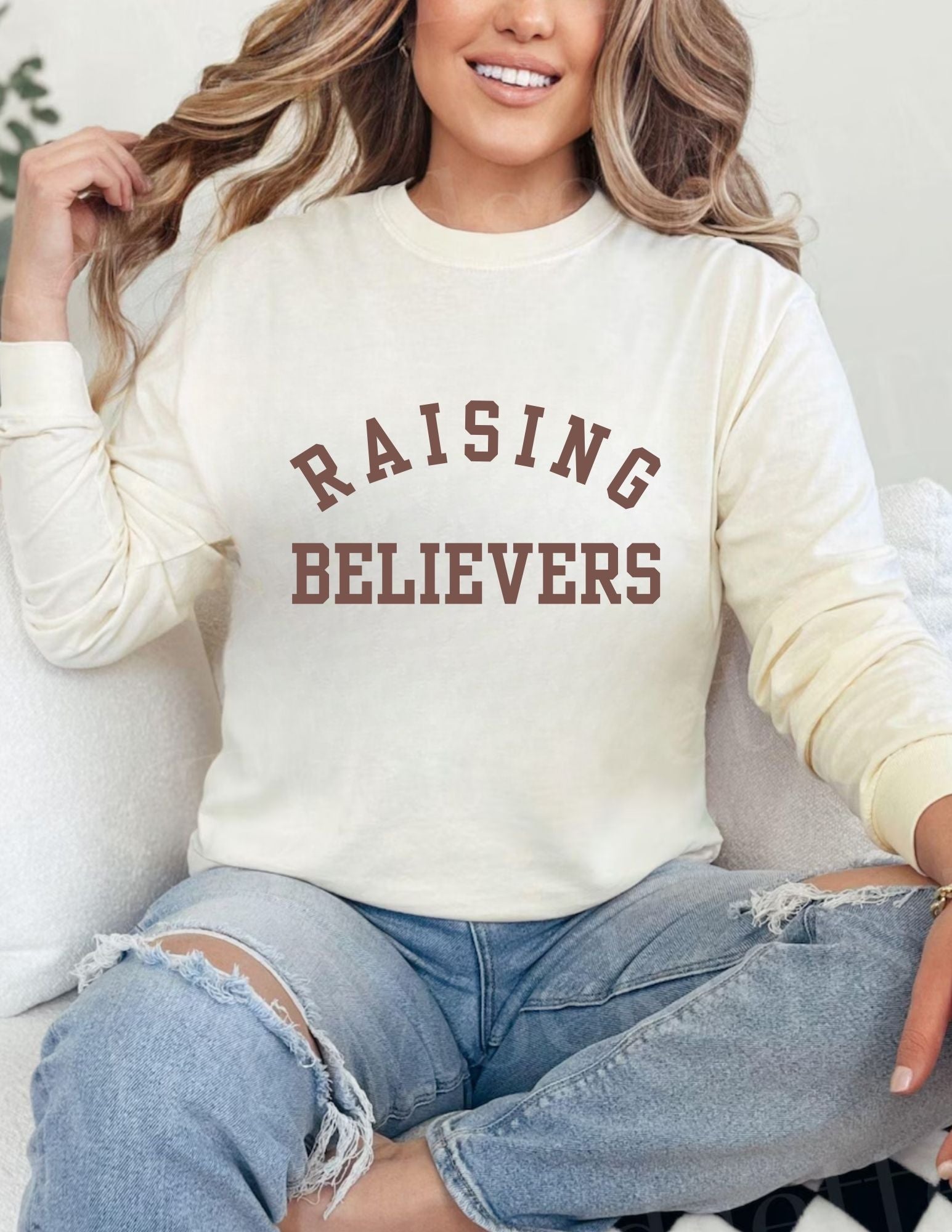Person wearing a cream sweatshirt with 'RAISING BELIEVERS' text, sitting on a couch.