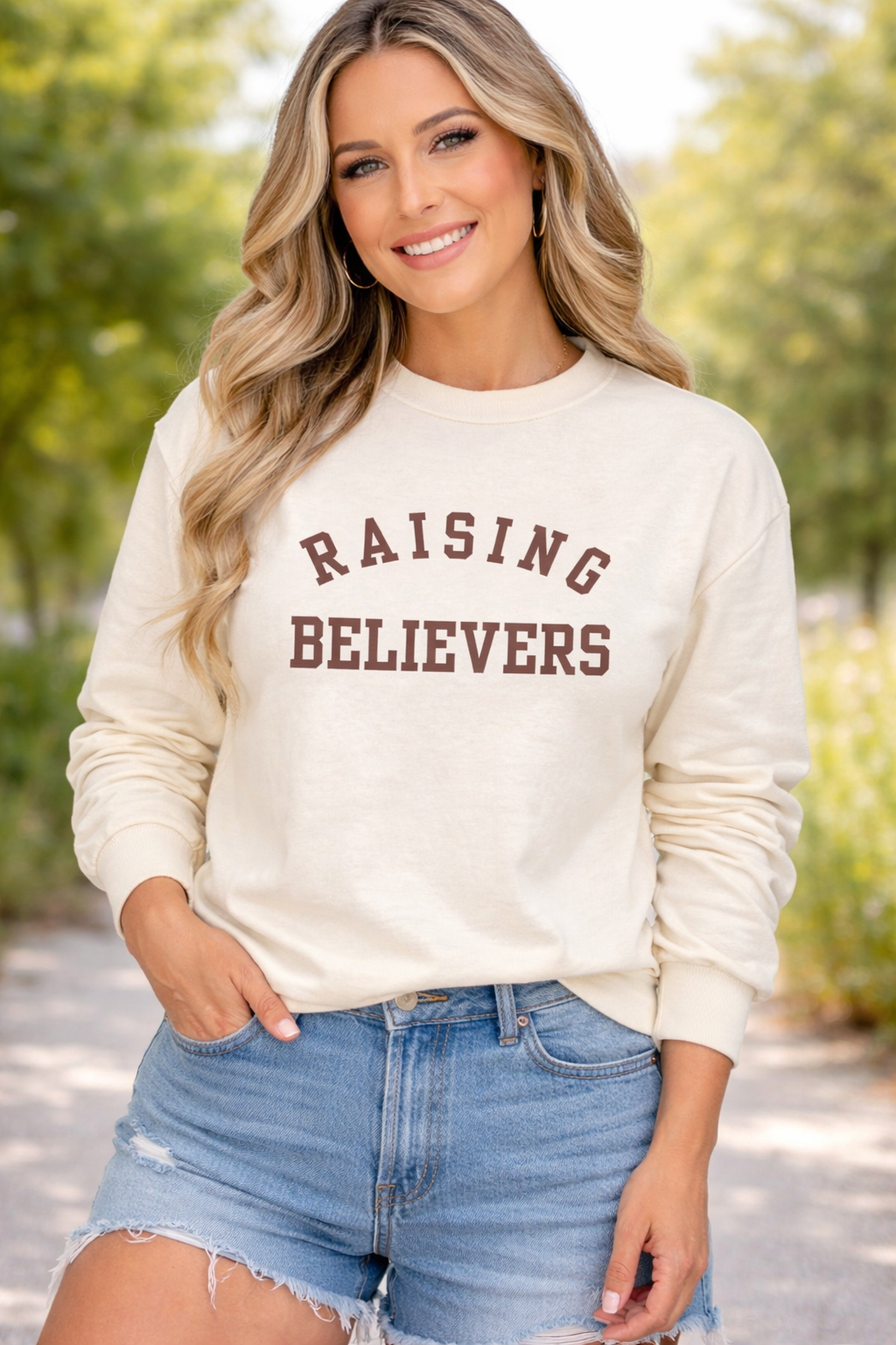 Woman wearing a cream sweatshirt with 'RAISING BELIEVERS' text, standing outdoors.