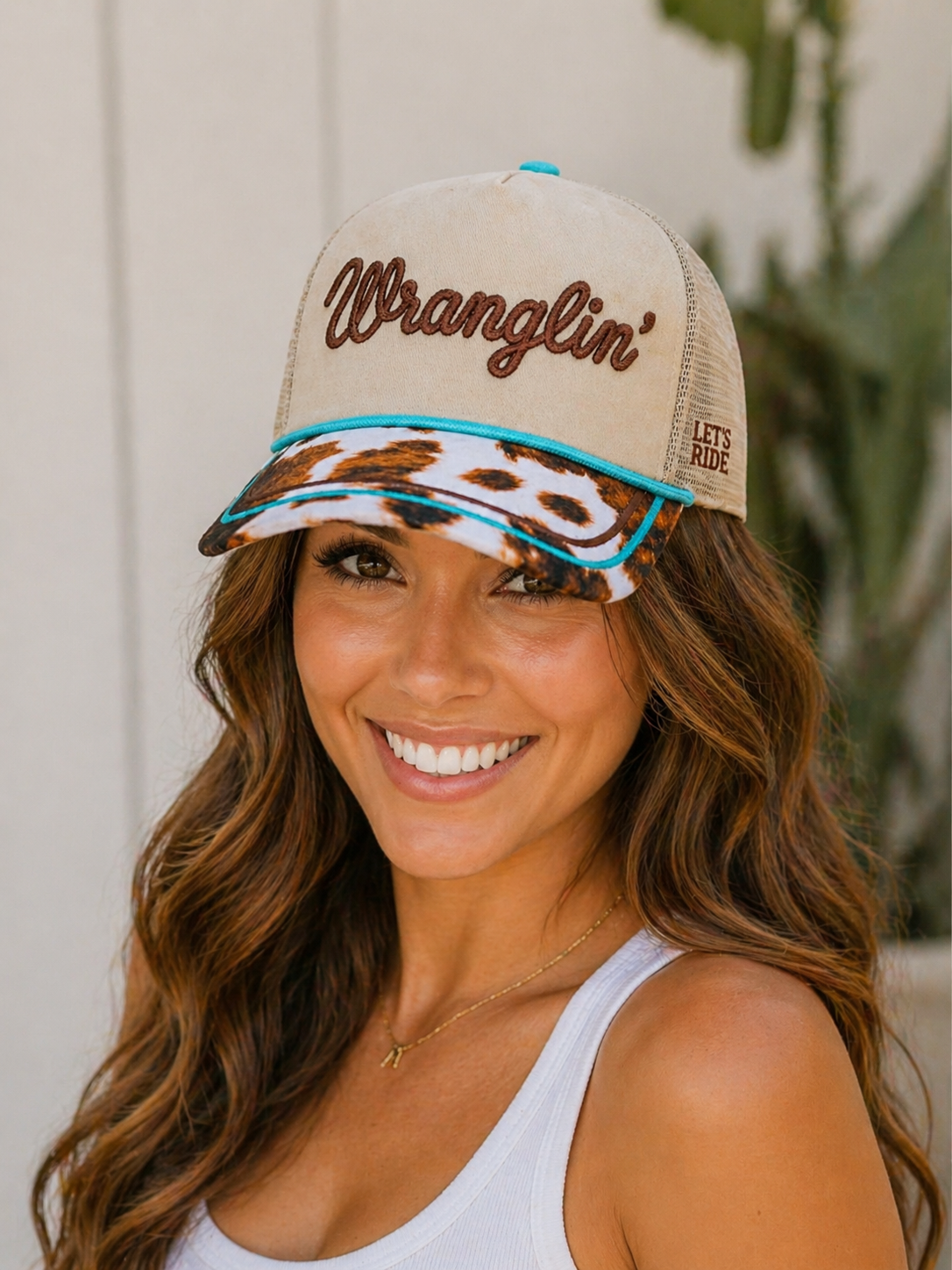 Woman wearing a beige cap with 'Wranglin'' text and a cow print design, smiling outdoors.